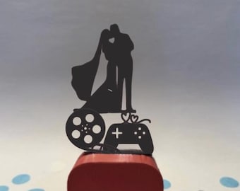 May include: A black silhouette wedding cake topper featuring a kissing couple, a film reel, and a video game controller. The topper sits atop a red square base, with blue and white confetti scattered around.