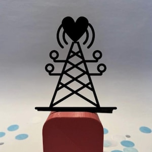 Peut inclure: Une tour métallique noire surmontée d'un cœur, posée sur un bloc rouge. La tour présente un motif en treillis et des éléments en forme d'antenne. Le cœur a un symbole de signal. Le bloc est de forme rectangulaire.