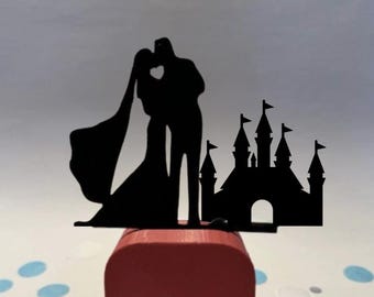 May include: A black silhouette wedding cake topper featuring a kissing couple with a heart-shaped cutout and a castle. The topper is mounted on a red square base, with blue confetti scattered around.