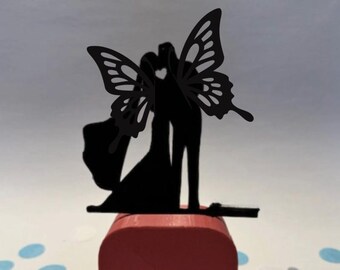 May include: A black silhouette of a couple embracing, with a butterfly wing design, atop a red wooden block. The design is likely a cake topper or decorative element for a wedding or special event.