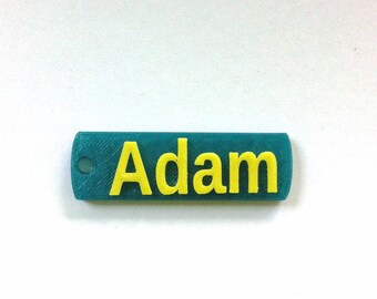 May include: A teal blue plastic name tag with a yellow inscription that reads "Adam".