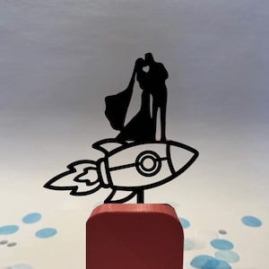 May include: A black silhouette wedding cake topper featuring a couple kissing atop a rocket ship. The rocket is white with black outlines and sits on a red square base. The background is a light gray with blue and white confetti.