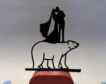 May include: A black silhouette wedding cake topper featuring a couple kissing atop a polar bear. The bear is white, and the topper is mounted on a red block. Blue and white confetti dots the background.