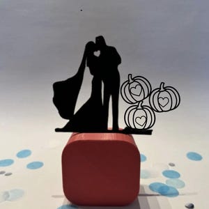May include: A black silhouette cake topper featuring a kissing couple with a heart-shaped cutout. Three pumpkin silhouettes with heart details are also included. The topper is mounted on a red square base, with blue and white confetti.