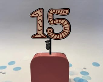 May include: A brown and beige number 15 cake topper with a small koala bear attached. The number is on a black stick, set in a red square base. Blue and white confetti dots the white surface.
