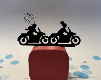May include: Black silhouette cake topper featuring a bride and groom riding motorcycles. The bride wears a veil, and the groom wears a top hat. The topper is placed on a red square base, with blue and white confetti in the background.