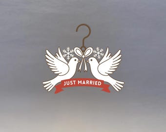 May include: A decorative wedding ornament featuring two white doves facing each other, connected by a bow and snowflakes. A red banner below reads "JUST MARRIED". The ornament hangs from a brown hanger.