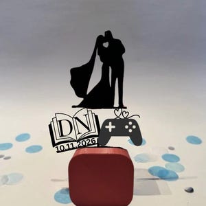 May include: A black wedding cake topper featuring a bride and groom silhouette kissing, with a heart-shaped cutout. Includes a book with initials "DN" and the date "10.11.2026", and a game controller. The topper sits on a red base.