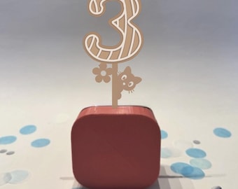 May include: A wooden number 3 cake topper with a light brown and white design. The number is on a stick with a cat and flower design. The topper is placed on a red square base. Confetti dots are scattered around the base.