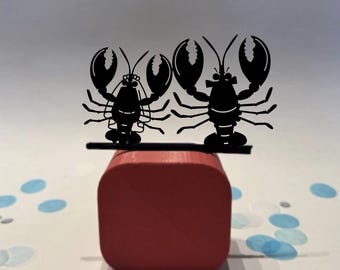 May include: Two black silhouette lobster decorations sit atop a small, red, rectangular block. The lobsters have large claws and detailed bodies. Blue confetti dots the white surface around the block, creating a playful scene.