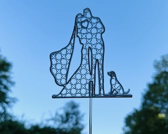 May include: A silver wedding cake topper featuring a bride and groom silhouette with a dog. The bride's veil has a decorative pattern. The topper is on a clear stick, perfect for a wedding cake.