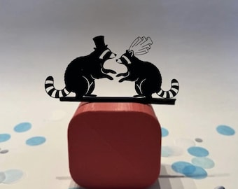May include: Black raccoon figurines, one wearing a top hat and the other a veil, stand on a black bar atop a red block. The raccoon figures are facing each other. The background is white with blue confetti.