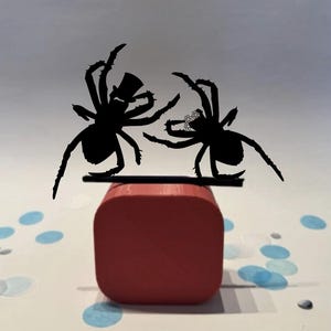 Puede incluir: Siluetas negras de dos arañas, una con sombrero de copa y otra con corona floral, sobre una plataforma negra encima de un cubo rojo. Fondo blanco con confeti azul. Un artículo decorativo.