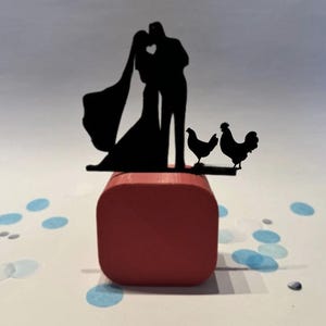 May include: A black silhouette wedding cake topper featuring a kissing couple with a bride's veil and two chickens. The topper is placed on a red square base, with blue confetti scattered around.