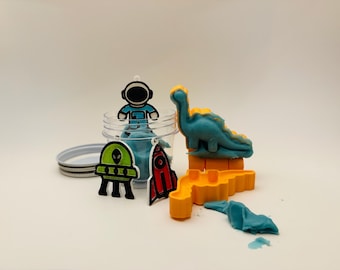 May include: A jar of blue playdough with space-themed cutouts and a dinosaur mold. The cutouts include an astronaut, a UFO, and a rocket. The dinosaur is teal with yellow accents.