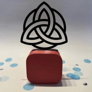Op de afbeelding: Een rode stempel met een zwart Keltisch knoopontwerp. De stempel is vierkant en heeft een zwart inktkussen. De Keltische knoop is een zwart, driedelig ontwerp. De achtergrond is wit met blauwe confetti.