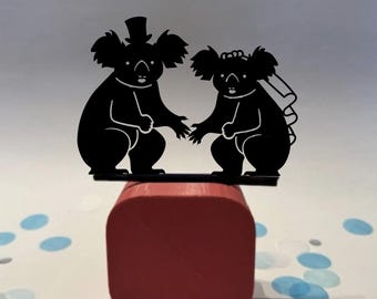 May include: A black silhouette of two koalas, one wearing a top hat and the other a veil, holding hands. The koalas are on a black platform atop a red wooden block. Blue and white confetti dots the background.