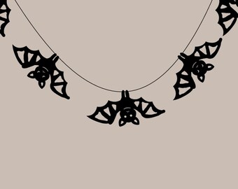 May include: A string of black bat cutouts hanging from a thin string against a neutral background. The bats are stylized with detailed wings and smiling faces, perfect for Halloween decorations.