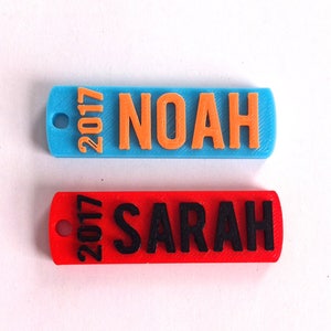 Puede incluir: Dos llaveros personalizados, uno azul con el nombre "NOAH" y el año "2017" en naranja, y otro rojo con el nombre "SARAH" y el año "2017" en negro.