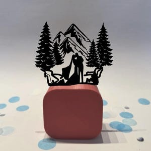 May include: A black silhouette wedding cake topper featuring a couple kissing in front of a mountain range and pine trees. The topper is mounted on a red square base, with blue and white confetti scattered around.