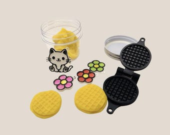 May include: A miniature black waffle iron sits open next to two yellow waffles. A clear jar with yellow contents, a cat charm, and flower charms are also present. The items are arranged on a light beige surface.
