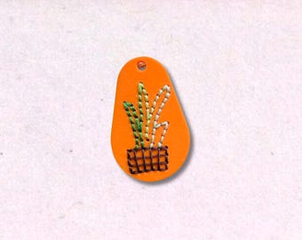 May include: Orange leather tag with a stitched plant design. The plant is in a black grid pot, with green, white, and yellow stitched leaves. The tag has a small hole at the top.