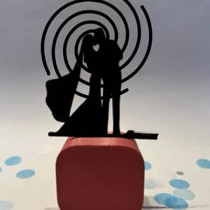 Op de afbeelding: Een zwarte silhouet bruidstaarttopper met een omhelzend stel, bovenop een rode vierkante basis. Een spiraalontwerp bevindt zich achter het paar. Blauwe confetti bedekt het witte oppervlak.