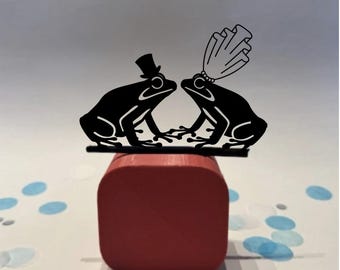 May include: A black metal wedding cake topper featuring two stylized frogs. One frog wears a top hat, the other a bridal veil. The topper sits atop a red square box, with blue and white confetti scattered around.