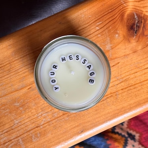 May include: A candle in a clear glass jar with the words "YOUR MESSAGE" spelled out in white letter beads. The candle has a light yellow hue and sits on a wooden surface.