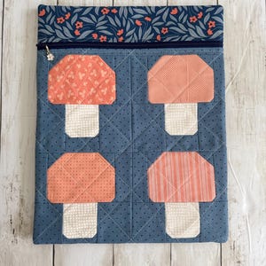 May include: A rectangular fabric pouch featuring a blue background with a dark blue zipper closure. The front displays four quilted mushroom shapes in varying shades of orange and cream. The top border has a floral pattern.