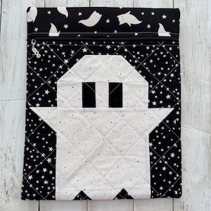 May include: A rectangular quilted pouch featuring a black fabric background with white stars and a central white ghost applique with black rectangular eyes. The top section has a zipper closure with a metal charm labeled moo.