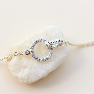 Op de afbeelding: Een delicate zilveren ketting met twee in elkaar grijpende ringen. Eén ring is bezet met kleine, heldere kristallen, de andere draagt de naam "Anna". De ketting ligt op een witte steen tegen een zachte, neutrale achtergrond.