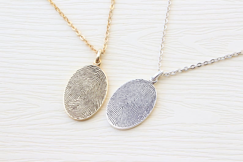 May include: Two silver necklaces with oval pendants featuring a fingerprint design. One necklace has a gold chain and pendant, while the other has a silver chain and pendant.