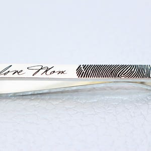 May include: A silver tie clip featuring a fingerprint pattern and the handwritten text "love Mom". The clip has a curved design. The background is a light, neutral colour.