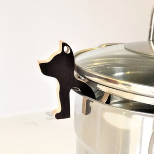 Könnte beinhalten: Ein schwarzer und hölzerner Topfdeckelhalter in Katzenform. Der Halter ist am Deckel eines Topfes aus Edelstahl befestigt.