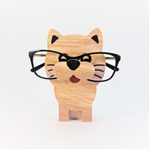 Puede incluir: Portagafas de madera con forma de gato con gafas negras en la parte superior. El gato tiene una cara pintada de marrón y negro con una lengua roja.