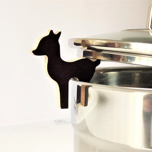 Puede incluir: Un soporte de tapa de olla de madera negra en forma de silueta de ciervo. El soporte está unido a una olla de acero inoxidable con tapa.