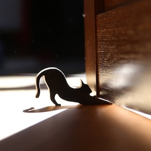 Könnte beinhalten: Ein schwarzer Katzensilhouetten-Türstopper, an einer Holztür platziert, mit einem Lichtstrahl, der auf den Boden fällt.