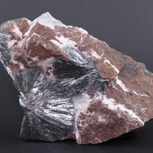 May include: A close-up of a silver-colored mineral specimen embedded in a reddish-brown rock matrix. The mineral has a radiating, starburst-like pattern.