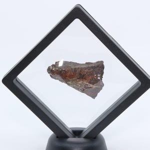 May include: A rough, dark brown and reddish-brown mineral specimen suspended within a square, black floating display frame. The frame is positioned at a diamond angle and is supported by a circular black base, creating a clear, hovering effect.