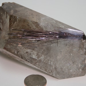 May include: A large, clear quartz crystal with a cluster of black tourmaline inclusions. The crystal is faceted and has a pointed tip. The crystal is approximately 12.5 cm long and 5 cm wide.