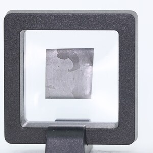 May include: A small, rectangular piece of a silvery-gray metal is displayed in a clear plastic frame. The metal has a rough, textured surface.