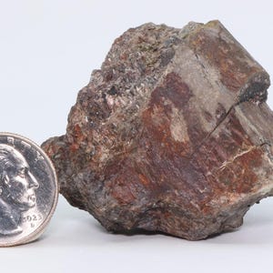 May include: A rough, irregularly shaped mineral specimen with a mix of reddish-brown, gray, and metallic dark tones. The rock features a crystalline texture and is positioned next to a silver-colored United States quarter coin for scale.