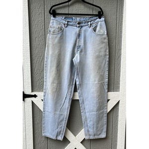 Pode incluir: Calças de ganga azul claro penduradas num cabide preto. As calças de ganga têm um design clássico de cinco bolsos, fecho de botão e zíper e um corte de perna reto. A ganga tem uma lavagem desbotada, dando-lhe um aspeto vintage.
