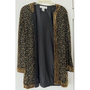 May include: A black and gold sequined jacket with a long sleeve and a front closure. The jacket has a subtle sheen and a delicate pattern.