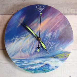 Puede incluir: Un reloj redondo de madera con un paisaje marítimo tormentoso azul, morado y amarillo. La esfera del reloj tiene agujas negras y un centro dorado. El reloj tiene las palabras "All Storms Pass" en forma de corazón en la parte superior.