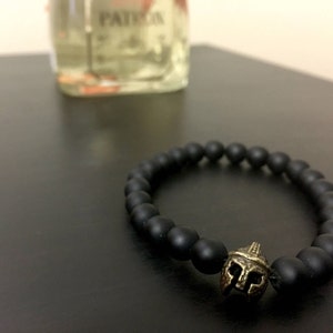 May include: Black beaded bracelet with a bronze-colored Spartan helmet charm. The bracelet is made of matte black beads and is displayed on a dark surface. A bottle is blurred in the background.