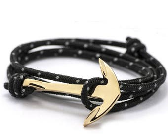 May include: A bracelet featuring a polished gold-tone metal anchor charm attached to a black cord. The cord is woven with small, reflective silver-coloured flecks throughout its length and is tied in a knot to secure the anchor in place.