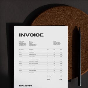 May include: A white invoice with black text, titled "INVOICE," lies on a dark surface. A black pencil rests beside the invoice. The invoice details services, quantities, and rates. A brown, circular coaster is in the background.
