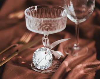 May include: A white round thank you tag with black floral illustrations and the text "Thank you for celebrating with us Joseph and Victoria" tied to a crystal glass with a white ribbon.